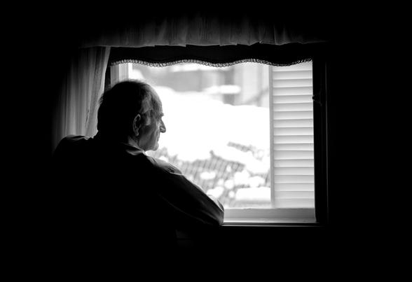 Black and white image of a lonely man in a dark room looking out a window