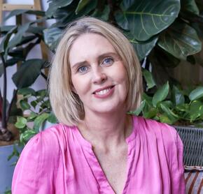 Image of Bretine Curtis, sitting, smiling at camera. Bretine is wearing a pink satin blouse and has shoulder lengthen blonde hair and blue eyes.